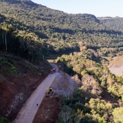A fotografia em plano panor&acirc;mico e &acirc;ngulo elevado captura uma estrada sinuosa que contorna a encosta de um morro, margeando um rio caudaloso em uma vasta regi&atilde;o montanhosa e vales verdejantes.

A Rodovia e as Obras (Centro-Esquerda):

Uma estrada estreita de asfalto claro corta a imagem diagonalmente a partir do canto inferior esquerdo em dire&ccedil;&atilde;o ao centro.

&Agrave; esquerda da pista, a encosta do morro apresenta trechos de terra roxa e encostas escavadas, evidenciando marcas de deslizamentos anteriores ou trabalhos de terraplanagem. Cones de sinaliza&ccedil;&atilde;o vermelhos e brancos est&atilde;o perfilados na margem para isolar a pista.

No trecho inferior da estrada, uma pequena escavadeira amarela est&aacute; posicionada na margem direita da via, onde h&aacute; um ac&uacute;mulo de terra batida e pedras. Pr&oacute;ximo a ela, duas pessoas caminham a p&eacute;. Mais &agrave; frente, na curva da estrada, um caminh&atilde;o de carga de cor vermelha segue viagem.

O Rio e o Vale (Direita): Correndo paralelamente &agrave; rodovia, um grande rio de &aacute;guas barrentas em tom marrom-claro serpenteia pelo vale &agrave; direita. Suas margens s&atilde;o irregulares e cobertas por pedregais e vegeta&ccedil;&atilde;o rasteira. Ao longe, no canto superior direito, nota-se uma ponte que cruza o leito do rio.

A Paisagem e o Relevo:

A paisagem &eacute; imponente e dominada por grandes montanhas e morros recobertos por uma floresta densa e cont&iacute;nua de Mata Atl&acirc;ntica em tons de verde-escuro.

A ilumina&ccedil;&atilde;o solar vem da direita, projetando sombras longas das &aacute;rvores e do pr&oacute;prio relevo sobre a encosta e a estrada no lado esquerdo, sugerindo o in&iacute;cio da manh&atilde; ou o final da tarde. O c&eacute;u na parte superior est&aacute; limpo e claro.

A imagem registra a complexidade geogr&aacute;fica e os desafios de infraestrutura e engenharia para a manuten&ccedil;&atilde;o de rodovias em regi&otilde;es de serra.