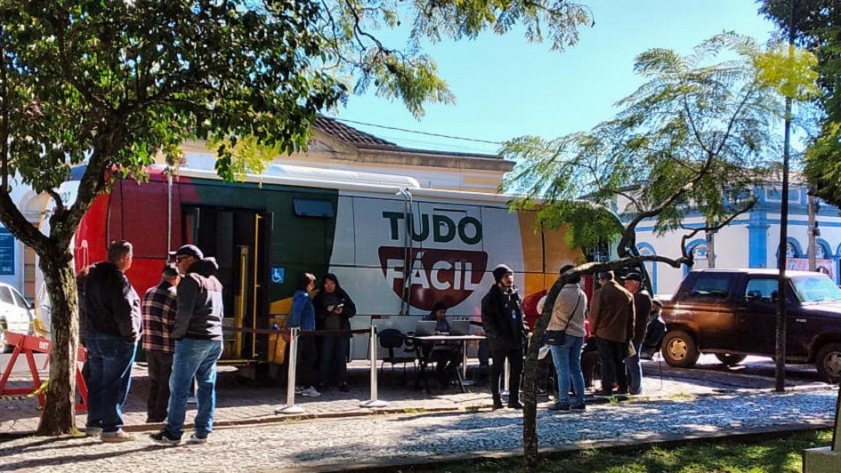 &Aacute;rea externa urbana com uma unidade m&oacute;vel identificada pelo texto &ldquo;TUDO F&Aacute;CIL&rdquo; estacionada junto &agrave; cal&ccedil;ada, onde v&aacute;rias pessoas est&atilde;o reunidas formando uma pequena fila pr&oacute;xima &agrave; entrada.