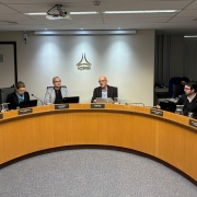 A fotografia em plano geral e &acirc;ngulo frontal mostra uma sess&atilde;o de julgamento ou reuni&atilde;o deliberativa institucional ocorrendo em um plen&aacute;rio de formato semicircular.

A Mesa Diretora: No centro do quadro, seis conselheiros (uma mulher e cinco homens) est&atilde;o sentados em cadeiras de escrit&oacute;rio pretas de encosto alto, acomodados atr&aacute;s de uma grande bancada curva de madeira em tom castanho-claro.

Os Integrantes (da esquerda para a direita):

Na extremidade esquerda, um senhor calvo de palet&oacute; escuro e camisa verde analisa pap&eacute;is em suas m&atilde;os. Ladeando-o, no canto esquerdo, est&atilde;o dispostas as bandeiras oficiais do Brasil e do Estado do Rio Grande do Sul em seus mastros.

Ao lado dele, uma mulher usa &oacute;culos e blusa escura com len&ccedil;o azul-claro no pesco&ccedil;o, concentrada diante de um notebook preto aberto.

No centro-esquerda, um homem de &oacute;culos e blazer cinza claro sobre camiseta escura observa a sess&atilde;o.

No centro da mesa, um homem calvo e de &oacute;culos, vestindo palet&oacute; escuro sobre camisa azul-clara, est&aacute; com as m&atilde;os gesticuladas e a boca aberta, indicando o uso da palavra. Ele tem um notebook prateado &agrave; sua frente.

&Agrave; sua direita (direita da foto), um jovem de barba, &oacute;culos e terno escuro sobre camisa clara acompanha a delibera&ccedil;&atilde;o.

Na extremidade direita, um senhor calvo e de barba, vestindo uma camisa social branca com as mangas dobradas, manipula documentos impressos sobre a bancada.

O Ambiente:

O piso do plen&aacute;rio &eacute; revestido por um carpete cinza-escuro texturizado. Na borda superior de cada assento da bancada, h&aacute; pequenas placas de identifica&ccedil;&atilde;o pretas com letras douradas indicando os nomes e cargos dos conselheiros. Sobre a mesa, h&aacute; notebooks, microfones de haste flex&iacute;vel e jarras de vidro com &aacute;gua.

A parede de fundo &eacute; de cor clara e exibe, centralizado logo acima do presidente da sess&atilde;o, o logotipo met&aacute;lico dourado da AGERGS (Ag&ecirc;ncia Estadual de Regula&ccedil;&atilde;o dos Servi&ccedil;os P&uacute;blicos Delegados do Rio Grande do Sul), composto por tr&ecirc;s linhas curvas que convergem no topo sobre a inscri&ccedil;&atilde;o do nome da ag&ecirc;ncia.

O teto &eacute; composto por placas modulares brancas com lumin&aacute;rias embutidas e um projetor de v&iacute;deo fixado ao centro. No canto superior direito, cortinas verticais claras cobrem as janelas.

A imagem registra uma reuni&atilde;o t&eacute;cnica e oficial de regula&ccedil;&atilde;o p&uacute;blica e tomada de decis&otilde;es administrativas.