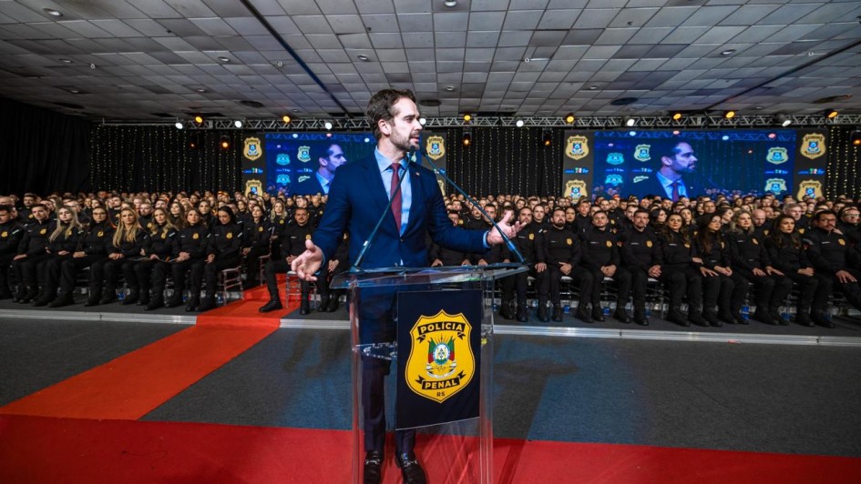 O governador discursa em p&uacute;lpito transparente com o bras&atilde;o &ldquo;Pol&iacute;cia Penal RS&rdquo;, diante de uma plateia extensa de participantes uniformizados, com tel&otilde;es replicando a cena e ilumina&ccedil;&atilde;o c&ecirc;nica no ambiente.