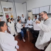 Fotografia mostra um grupo de cerca de 12 pessoas usando jalecos brancos reunidas em um laborat&oacute;rio. Em primeiro plano, um homem de jaleco gesticula enquanto segura folhas de papel e fala aos participantes, que o observam atentamente. Ao redor, alguns integrantes est&atilde;o em p&eacute; e outros sentados junto a uma bancada central. O ambiente cont&eacute;m equipamentos laboratoriais, arm&aacute;rios suspensos, pipetas, suportes para tubos de ensaio e outros materiais de pesquisa, indicando uma atividade de treinamento ou orienta&ccedil;&atilde;o t&eacute;cnica.