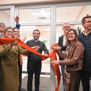 Grupo de pessoas em ambiente interno corta uma fita vermelha na entrada de um espa&ccedil;o com portas de vidro, marcando uma inaugura&ccedil;&atilde;o; o local tem ilumina&ccedil;&atilde;o clara e paredes neutras.
