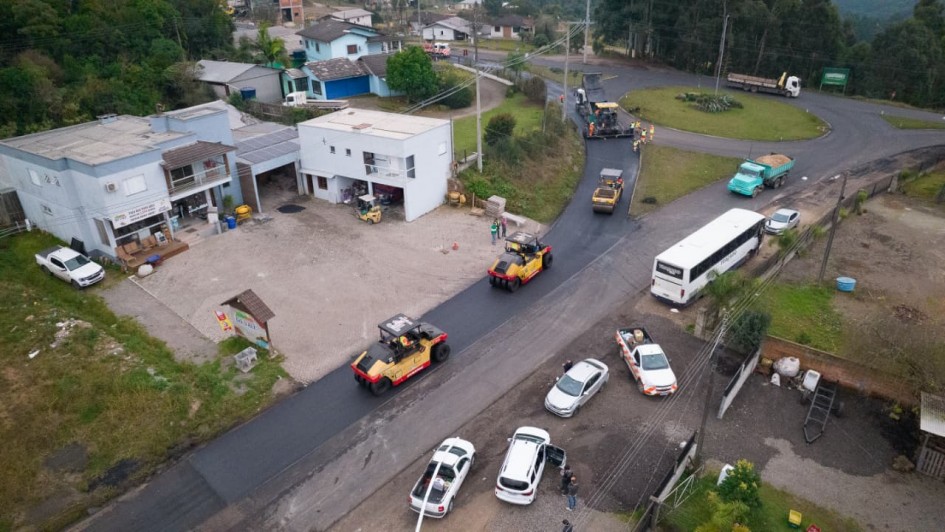 Vista a&eacute;rea de estrada em &aacute;rea semiurbana com m&aacute;quinas realizando asfaltamento pr&oacute;ximo a uma rotat&oacute;ria, ve&iacute;culos estacionados nas margens e resid&ecirc;ncias ao redor.