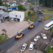 Vista a&eacute;rea de estrada em &aacute;rea semiurbana com m&aacute;quinas realizando asfaltamento pr&oacute;ximo a uma rotat&oacute;ria, ve&iacute;culos estacionados nas margens e resid&ecirc;ncias ao redor.