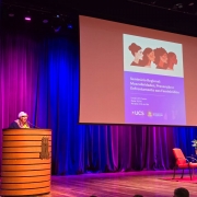 A fotografia registra a realiza&ccedil;&atilde;o de um evento institucional em um audit&oacute;rio, identificado pelo nome do arquivo e pelo slide projetado como o "Semin&aacute;rio Regional: Masculinidades, Preven&ccedil;&atilde;o e Enfrentamento aos Feminic&iacute;dios".

O Palco e a Palestrante
A&ccedil;&atilde;o Principal: No lado esquerdo, uma mulher de cabelos curtos e claros, usando &oacute;culos de arma&ccedil;&atilde;o vermelha e um len&ccedil;o colorido, fala ao microfone posicionada atr&aacute;s de um p&uacute;lpito cil&iacute;ndrico de madeira com detalhes em frisos horizontais.

Mobili&aacute;rio do Palco: No lado direito do palco, sobre um tapete vermelho, h&aacute; duas poltronas estofadas tamb&eacute;m na cor vermelha e uma pequena mesa de centro de vidro com um arranjo de flores (ant&uacute;rios), configurando um espa&ccedil;o para debates.

Elementos de Proje&ccedil;&atilde;o
Tel&atilde;o Central: Uma grande tela de proje&ccedil;&atilde;o est&aacute; centralizada no palco, exibindo a identidade visual do evento. Na parte superior do slide, h&aacute; uma ilustra&ccedil;&atilde;o estilizada com o perfil de quatro mulheres de diferentes etnias.

Texto do Slide: Logo abaixo da ilustra&ccedil;&atilde;o, o slide em tom lil&aacute;s traz o t&iacute;tulo do semin&aacute;rio e informa&ccedil;&otilde;es institucionais, incluindo o local (UCS Teatro), a data (10/06) e o hor&aacute;rio (09h &agrave;s 18h). No rodap&eacute; do slide, destacam-se os logotipos da UCS (Universidade de Caxias do Sul) e do Governo do Estado do Rio Grande do Sul.

Ambiente e Ilumina&ccedil;&atilde;o
Cen&aacute;rio: O fundo do palco &eacute; composto por grandes cortinas de tecido que recebem uma ilumina&ccedil;&atilde;o c&ecirc;nica marcante em tons de azul e violeta. A ilumina&ccedil;&atilde;o cria um forte contraste com o piso de madeira do palco e a estrutura c&ecirc;nica superior.

Perspectiva: A foto foi tirada a partir da plateia, ligeiramente de baixo para cima, mostrando o topo da cabe&ccedil;a de um espectador em primeiro plano no canto inferior direito.