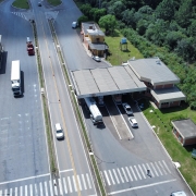 Esta &eacute; uma descri&ccedil;&atilde;o da imagem para fins de acessibilidade:

Uma fotografia a&eacute;rea, capturada por um drone em &acirc;ngulo picado (de cima para baixo), mostra a estrutura do Posto Fiscal de Vacaria, localizado &agrave;s margens de uma rodovia duplicada.

Estrutura Principal e Rodovia
O Posto Fiscal: No centro da imagem, destaca-se uma grande cobertura met&aacute;lica arqueada e alongada, pintada de branco com detalhes em azul nas bordas frontais. Sob essa estrutura, encontram-se as pistas de triagem e pesagem para os ve&iacute;culos de carga.

A Rodovia: A estrada possui pistas duplicadas em asfalto escuro, separadas por uma barreira central de concreto (tipo New Jersey) e faixas sinalizadoras pintadas em amarelo e branco. O fluxo de ve&iacute;culos &eacute; moderado, observando-se alguns caminh&otilde;es e carros transitando ou estacionados nas proximidades do posto.

Edifica&ccedil;&otilde;es de Apoio: &Agrave; esquerda da grande cobertura, h&aacute; pr&eacute;dios administrativos de apoio com telhados em tons de cinza e &aacute;reas de estacionamento interno para ve&iacute;culos oficiais e de funcion&aacute;rios.

Ambiente e Entorno
Vegeta&ccedil;&atilde;o: A infraestrutura rodovi&aacute;ria &eacute; cercada por amplas &aacute;reas verdes. H&aacute; gramados bem cuidados nas margens e canteiros, al&eacute;m de copas de &aacute;rvores nativas dispersas ao redor do complexo.

Ilumina&ccedil;&atilde;o e Clima: A imagem foi registrada em um dia claro, com luz solar direta que projeta sombras n&iacute;tidas das &aacute;rvores, dos ve&iacute;culos e da pr&oacute;pria estrutura met&aacute;lica sobre o solo. Ao fundo, a paisagem se estende em &aacute;reas de campo aberto sob uma linha de horizonte suave.