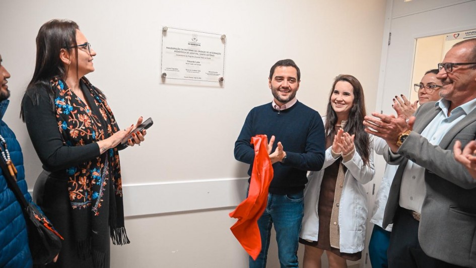 Grupo posa para a foto batendo palmas em frente a uma placa de inaugura&ccedil;&atilde;o fixada na parede. 