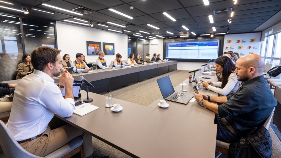 A fotografia em plano geral e &acirc;ngulo diagonal registra uma reuni&atilde;o t&eacute;cnica de trabalho em uma sala de confer&ecirc;ncias moderna, identificada pelo t&iacute;tulo do arquivo como o an&uacute;ncio de um semin&aacute;rio estadual de prepara&ccedil;&atilde;o ao fen&ocirc;meno clim&aacute;tico El Ni&ntilde;o.

Disposi&ccedil;&atilde;o e Participantes
A Grande Mesa: Os participantes (mais de 15 pessoas) est&atilde;o sentados ao redor de uma grande mesa cinza em formato de "U". A imagem &eacute; capturada a partir de uma das extremidades abertas da mesa, criando uma perspectiva que alonga a sala.

Primeiro Plano: &Agrave; esquerda, em destaque pr&oacute;ximo &agrave; c&acirc;mera, um homem com barba e camisa social branca est&aacute; sentado de perfil, com as m&atilde;os juntas pr&oacute;ximas ao rosto, observando a reuni&atilde;o. &Agrave; direita, um homem careca com camisa preta mexe em um smartphone. Ao lado dele, uma mulher de blusa branca tamb&eacute;m interage com um dispositivo m&oacute;vel.

Equipes de Apoio: Ao longo do bra&ccedil;o esquerdo da mesa, veem-se integrantes vestindo coletes oficiais bicolores (laranja na parte superior e azul-marinho na inferior), caracter&iacute;sticos da Defesa Civil. Outros participantes em trajes casuais de neg&oacute;cios completam a mesa.

Objetos sobre a Mesa: Est&atilde;o distribu&iacute;dos notebooks abertos, tablets, microfones de mesa com pedestais pretos, copos de &aacute;gua, x&iacute;caras de caf&eacute; e blocos de papel com canetas.

O Ambiente e Recursos Visuais
Painel Digital: Na parede ao fundo, centralizada, h&aacute; uma grande tela que projeta um sistema com dados e tabelas estruturadas em tons de azul e branco.

Identidade Visual: &Agrave; direita da tela, um painel vertical branco exibe logotipos coloridos repetidos com a inscri&ccedil;&atilde;o "Rio Grande do Sul", identificando o governo do estado. Na parede lateral esquerda, h&aacute; dois quadros decorativos pequenos e horizontais com imagens coloridas.

Infraestrutura: A sala tem teto preto texturizado com lumin&aacute;rias de LED retangulares embutidas e trilhos de spots direcionais acesos. O piso &eacute; revestido com carpete cinza-claro e, &agrave; extrema direita, h&aacute; grandes janelas de vidro que cobrem a parede lateral.