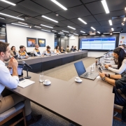 A fotografia em plano geral e &acirc;ngulo diagonal registra uma reuni&atilde;o t&eacute;cnica de trabalho em uma sala de confer&ecirc;ncias moderna, identificada pelo t&iacute;tulo do arquivo como o an&uacute;ncio de um semin&aacute;rio estadual de prepara&ccedil;&atilde;o ao fen&ocirc;meno clim&aacute;tico El Ni&ntilde;o.

Disposi&ccedil;&atilde;o e Participantes
A Grande Mesa: Os participantes (mais de 15 pessoas) est&atilde;o sentados ao redor de uma grande mesa cinza em formato de "U". A imagem &eacute; capturada a partir de uma das extremidades abertas da mesa, criando uma perspectiva que alonga a sala.

Primeiro Plano: &Agrave; esquerda, em destaque pr&oacute;ximo &agrave; c&acirc;mera, um homem com barba e camisa social branca est&aacute; sentado de perfil, com as m&atilde;os juntas pr&oacute;ximas ao rosto, observando a reuni&atilde;o. &Agrave; direita, um homem careca com camisa preta mexe em um smartphone. Ao lado dele, uma mulher de blusa branca tamb&eacute;m interage com um dispositivo m&oacute;vel.

Equipes de Apoio: Ao longo do bra&ccedil;o esquerdo da mesa, veem-se integrantes vestindo coletes oficiais bicolores (laranja na parte superior e azul-marinho na inferior), caracter&iacute;sticos da Defesa Civil. Outros participantes em trajes casuais de neg&oacute;cios completam a mesa.

Objetos sobre a Mesa: Est&atilde;o distribu&iacute;dos notebooks abertos, tablets, microfones de mesa com pedestais pretos, copos de &aacute;gua, x&iacute;caras de caf&eacute; e blocos de papel com canetas.

O Ambiente e Recursos Visuais
Painel Digital: Na parede ao fundo, centralizada, h&aacute; uma grande tela que projeta um sistema com dados e tabelas estruturadas em tons de azul e branco.

Identidade Visual: &Agrave; direita da tela, um painel vertical branco exibe logotipos coloridos repetidos com a inscri&ccedil;&atilde;o "Rio Grande do Sul", identificando o governo do estado. Na parede lateral esquerda, h&aacute; dois quadros decorativos pequenos e horizontais com imagens coloridas.

Infraestrutura: A sala tem teto preto texturizado com lumin&aacute;rias de LED retangulares embutidas e trilhos de spots direcionais acesos. O piso &eacute; revestido com carpete cinza-claro e, &agrave; extrema direita, h&aacute; grandes janelas de vidro que cobrem a parede lateral.