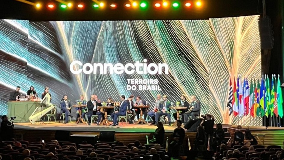 Foto mostra autoridades sentadas no palco, num cen&aacute;rio que lembra um restaurante, com um pequeno bar ao lado. No outro ponto do palco, v&aacute;rias bandeiras. No fundo, um tel&atilde;o como nome do evento.  