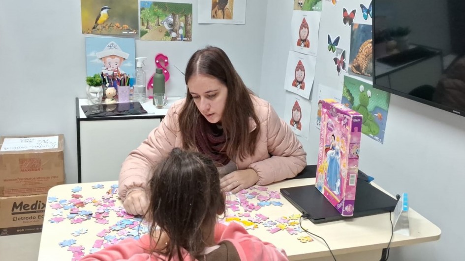 Sala de atendimento com decora&ccedil;&atilde;o infantil nas paredes, onde uma crian&ccedil;a e uma pessoa adulta montam um quebra-cabe&ccedil;a sobre uma mesa.