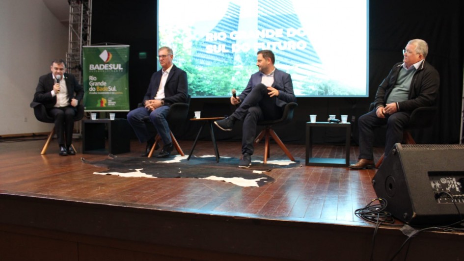 Esta &eacute; uma descri&ccedil;&atilde;o da imagem para fins de acessibilidade:

A fotografia em plano m&eacute;dio e &acirc;ngulo frontal registra um painel de debate ocorrendo no palco de um audit&oacute;rio, identificado pelo nome do arquivo como a participa&ccedil;&atilde;o do Governo do Estado no F&oacute;rum de Competitividade no Vale do Taquari.

Os Painelistas e a Disposi&ccedil;&atilde;o
Participantes: Quatro homens est&atilde;o sentados em poltronas pretas individuais com p&eacute;s de madeira, dispostas em linha ao longo do palco.

Composi&ccedil;&atilde;o (da esquerda para a direita):

O primeiro homem, vestindo terno escuro e camisa branca, est&aacute; ligeiramente inclinado para a frente e fala ao microfone.

O segundo homem usa um blazer escuro, camisa clara e cal&ccedil;a jeans, mantendo uma postura de escuta com as m&atilde;os cruzadas no colo.

O terceiro homem, com as pernas cruzadas, veste blazer escuro e cal&ccedil;a escura, segurando um microfone enquanto gesticula com a outra m&atilde;o.

O quarto homem, posicionado na extrema direita, usa &oacute;culos, uma blusa de gola alta clara sob uma jaqueta escura e cal&ccedil;a escura.

Elementos de Cenografia e Proje&ccedil;&atilde;o
Tel&atilde;o de Fundo: Uma grande tela digital centralizada ao fundo exibe uma imagem de um edif&iacute;cio moderno de arquitetura marcante e vegeta&ccedil;&atilde;o ao redor. Sobreposta &agrave; imagem, h&aacute; a inscri&ccedil;&atilde;o em letras mai&uacute;sculas brancas: "RIO GRANDE DO SUL DO FUTURO".

Banner Institucional: &Agrave; esquerda, h&aacute; um banner vertical verde com o logotipo e o nome do "BADESUL DESENVOLVIMENTO", seguido da frase "Rio Grande do Badesul".

Decora&ccedil;&atilde;o do Palco: O ch&atilde;o do palco &eacute; feito de t&aacute;buas de madeira compridas dispostas horizontalmente. No centro, sob as duas poltronas centrais, h&aacute; um grande tapete de couro bovino com manchas pretas e brancas. Pequenas mesas de apoio pretas com copos de &aacute;gua est&atilde;o posicionadas entre os participantes.

Ambiente e Detalhes T&eacute;cnicos
Estrutura: O palco &eacute; emoldurado por uma estrutura met&aacute;lica de treli&ccedil;a (box truss) vis&iacute;vel no canto superior esquerdo e por cortinas pretas ao fundo.

Equipamentos: No canto inferior direito, em primeiro plano, destaca-se uma caixa de retorno de som preta voltada para o palco, com alguns fios conectados.