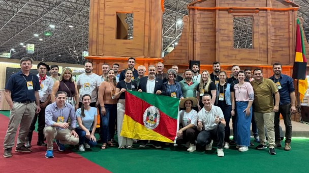 Foto mostra delega&ccedil;&atilde;o e feirantes reunidos para uma foto em grupo na frente de uma representa&ccedil;&atilde;o das ru&iacute;nas de S&atilde;o Miguel. O secret&aacute;rio Paim e outras pessoas seguram uma bandeira do Rio Grande do Sul.