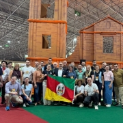 Foto mostra delega&ccedil;&atilde;o e feirantes reunidos para uma foto em grupo na frente de uma representa&ccedil;&atilde;o das ru&iacute;nas de S&atilde;o Miguel. O secret&aacute;rio Paim e outras pessoas seguram uma bandeira do Rio Grande do Sul.