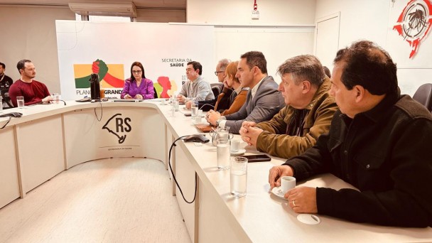 Ambiente interno de reuni&atilde;o institucional com mesa em formato curvo identificada com &ldquo;RS&rdquo; e &ldquo;Secretaria da Sa&uacute;de&rdquo;, onde v&aacute;rias pessoas est&atilde;o sentadas lado a lado participando de um encontro; ao fundo um painel exibe elementos gr&aacute;ficos e o texto &ldquo;Secretaria da Sa&uacute;de&rdquo; ao lado de um mapa do estado.