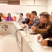 Ambiente interno de reuni&atilde;o institucional com mesa em formato curvo identificada com &ldquo;RS&rdquo; e &ldquo;Secretaria da Sa&uacute;de&rdquo;, onde v&aacute;rias pessoas est&atilde;o sentadas lado a lado participando de um encontro; ao fundo um painel exibe elementos gr&aacute;ficos e o texto &ldquo;Secretaria da Sa&uacute;de&rdquo; ao lado de um mapa do estado.