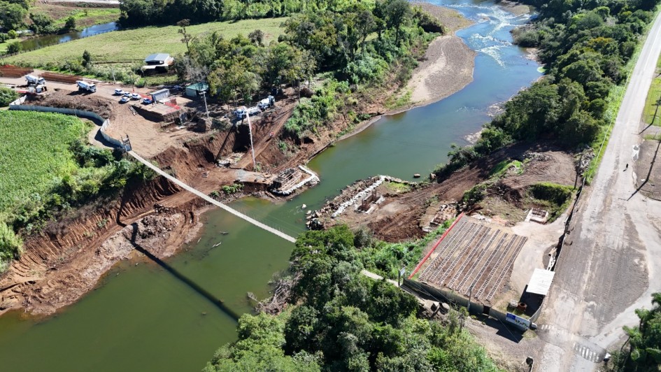 Vista a&eacute;rea de uma obra de ponte sobre um rio em &aacute;rea rural, com margens escavadas e estruturas em constru&ccedil;&atilde;o nas duas extremidades, al&eacute;m de m&aacute;quinas e ve&iacute;culos de apoio no canteiro.