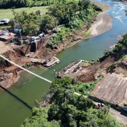 Vista a&eacute;rea de uma obra de ponte sobre um rio em &aacute;rea rural, com margens escavadas e estruturas em constru&ccedil;&atilde;o nas duas extremidades, al&eacute;m de m&aacute;quinas e ve&iacute;culos de apoio no canteiro.