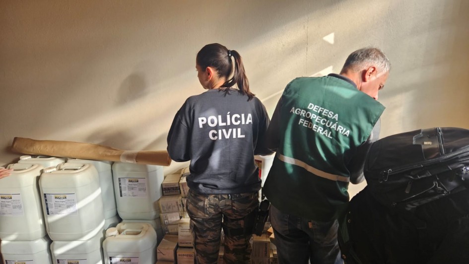 A fotografia em plano m&eacute;dio e &acirc;ngulo frontal registra uma a&ccedil;&atilde;o conjunta de fiscaliza&ccedil;&atilde;o e seguran&ccedil;a em um ambiente interno, identificada pelo t&iacute;tulo do arquivo como parte das a&ccedil;&otilde;es da Opera&ccedil;&atilde;o Pampa no combate a crimes rurais e transfronteiri&ccedil;os.

Agentes e A&ccedil;&atilde;o
Os Fiscais: Em primeiro plano, duas pessoas s&atilde;o vistas de costas enquanto examinam produtos armazenados.

Policial Civil: &Agrave; esquerda, uma mulher com o cabelo preso em um rabo de cavalo veste um moletom preto com a inscri&ccedil;&atilde;o "POL&Iacute;CIA CIVIL" em letras mai&uacute;sculas brancas nas costas. Ela usa cal&ccedil;a com estampa camuflada em tons de verde, marrom e preto.

Fiscal Federal: &Agrave; direita, um homem de cabelos curtos grisalhos veste um colete verde-escuro com uma faixa refletiva cinza horizontal. Nas costas do colete est&aacute; escrito, em letras mai&uacute;sculas brancas: "DEFESA AGROPECU&Aacute;RIA / FEDERAL".

Materiais e Ambiente
Produtos Apreendidos ou Fiscalizados: Diante dos agentes, h&aacute; uma pilha de insumos agr&iacute;colas. No lado esquerdo, observam-se diversos gal&otilde;es pl&aacute;sticos grandes e brancos empilhados (com r&oacute;tulos onde se l&ecirc; parcialmente a marca "MASTER"). No centro e ao fundo, h&aacute; caixas de papel&atilde;o empilhadas com inscri&ccedil;&otilde;es t&eacute;cnicas (onde se l&ecirc; "AGROPEC"). Um longo rolo de papel pardo est&aacute; apoiado horizontalmente sobre os gal&otilde;es.

Cen&aacute;rio e Ilumina&ccedil;&atilde;o: A a&ccedil;&atilde;o ocorre junto a uma parede lisa de tom claro (bege ou cinza-claro). Uma forte luz solar lateral e natural entra pelo lado direito, projetando feixes luminosos e sombras marcadas sobre a parede e as costas dos agentes. No canto inferior direito, v&ecirc;-se parte de uma mochila ou bolsa preta em primeiro plano.