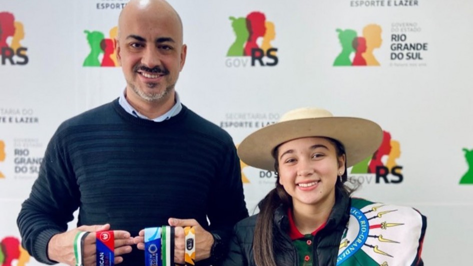 Foto mostra a atleta, com uma jaqueta escura e um chap&eacute;u aba larga claro, e com uma bandeira do Rio Grande do Sul sobre os ombros, segura uma medalha. Ao lado dela, o secret&aacute;rio Joel Maraschin, segura outras medalhas da atleta. 