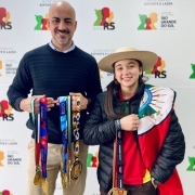 Foto mostra a atleta, com uma jaqueta escura e um chap&eacute;u aba larga claro, e com uma bandeira do Rio Grande do Sul sobre os ombros, segura uma medalha. Ao lado dela, o secret&aacute;rio Joel Maraschin, segura outras medalhas da atleta. 