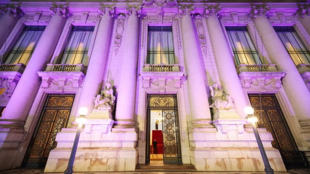 A fotografia em plano geral e &acirc;ngulo contrapicado (de baixo para cima) registra a imponente fachada do Pal&aacute;cio Piratini iluminada de forma tem&aacute;tica. O t&iacute;tulo do arquivo associa a a&ccedil;&atilde;o &agrave; celebra&ccedil;&atilde;o dos **25 anos do Teste do Pezinho** e ao refor&ccedil;o da import&acirc;ncia do diagn&oacute;stico precoce no SUS.

### **Arquitetura e Ilumina&ccedil;&atilde;o**

* **A Ilumina&ccedil;&atilde;o Tem&aacute;tica:** Toda a imponente estrutura de pedra da fachada hist&oacute;rica est&aacute; banhada por uma ilumina&ccedil;&atilde;o c&ecirc;nica noturna em tons de lil&aacute;s e roxo. A luz colore as grandes colunas, os relevos ornamentados e as sacadas do edif&iacute;cio.
* **Elementos da Fachada:** A composi&ccedil;&atilde;o destaca a simetria cl&aacute;ssica do pal&aacute;cio, com altas colunas cil&iacute;ndricas que sustentam a parte superior da estrutura, janelas verticais compridas protegidas por cortinas claras no andar superior, e portas de ferro trabalhado no t&eacute;rreo. Um grande bras&atilde;o esculpido em pedra encima a entrada principal.
* **Est&aacute;tuas e Adornos:** Ladeando o portal central, h&aacute; duas grandes esculturas de m&aacute;rmore claro representando figuras humanas sentadas sobre pedestais integrados &agrave; arquitetura.

### **Entrada Principal e Interior**

* **O Portal Central:** A grande porta principal de ferro e vidro est&aacute; parcialmente aberta. Atrav&eacute;s dela, &eacute; poss&iacute;vel vislumbrar o interior iluminado do pal&aacute;cio, revelando um lustre cl&aacute;ssico aceso e uma escadaria interna imponente revestida com um tapete vermelho vivo.
* **Seguran&ccedil;a:** Uma silhueta escura de um seguran&ccedil;a ou guarda em p&eacute; &eacute; vis&iacute;vel no interior da entrada, logo &agrave; frente dos primeiros degraus da escada.

### **Primeiro Plano**

* **Ilumina&ccedil;&atilde;o P&uacute;blica:** No n&iacute;vel da cal&ccedil;ada, em frente ao pr&eacute;dio, dois postes de ilumina&ccedil;&atilde;o de estilo antigo (lampi&otilde;es) com globos esf&eacute;ricos brancos acesos est&atilde;o posicionados um de cada lado, projetando uma luz amarelada e quente na base da fachada.