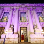 A fotografia em plano geral e &acirc;ngulo contrapicado (de baixo para cima) registra a imponente fachada do Pal&aacute;cio Piratini iluminada de forma tem&aacute;tica. O t&iacute;tulo do arquivo associa a a&ccedil;&atilde;o &agrave; celebra&ccedil;&atilde;o dos **25 anos do Teste do Pezinho** e ao refor&ccedil;o da import&acirc;ncia do diagn&oacute;stico precoce no SUS.

### **Arquitetura e Ilumina&ccedil;&atilde;o**

* **A Ilumina&ccedil;&atilde;o Tem&aacute;tica:** Toda a imponente estrutura de pedra da fachada hist&oacute;rica est&aacute; banhada por uma ilumina&ccedil;&atilde;o c&ecirc;nica noturna em tons de lil&aacute;s e roxo. A luz colore as grandes colunas, os relevos ornamentados e as sacadas do edif&iacute;cio.
* **Elementos da Fachada:** A composi&ccedil;&atilde;o destaca a simetria cl&aacute;ssica do pal&aacute;cio, com altas colunas cil&iacute;ndricas que sustentam a parte superior da estrutura, janelas verticais compridas protegidas por cortinas claras no andar superior, e portas de ferro trabalhado no t&eacute;rreo. Um grande bras&atilde;o esculpido em pedra encima a entrada principal.
* **Est&aacute;tuas e Adornos:** Ladeando o portal central, h&aacute; duas grandes esculturas de m&aacute;rmore claro representando figuras humanas sentadas sobre pedestais integrados &agrave; arquitetura.

### **Entrada Principal e Interior**

* **O Portal Central:** A grande porta principal de ferro e vidro est&aacute; parcialmente aberta. Atrav&eacute;s dela, &eacute; poss&iacute;vel vislumbrar o interior iluminado do pal&aacute;cio, revelando um lustre cl&aacute;ssico aceso e uma escadaria interna imponente revestida com um tapete vermelho vivo.
* **Seguran&ccedil;a:** Uma silhueta escura de um seguran&ccedil;a ou guarda em p&eacute; &eacute; vis&iacute;vel no interior da entrada, logo &agrave; frente dos primeiros degraus da escada.

### **Primeiro Plano**

* **Ilumina&ccedil;&atilde;o P&uacute;blica:** No n&iacute;vel da cal&ccedil;ada, em frente ao pr&eacute;dio, dois postes de ilumina&ccedil;&atilde;o de estilo antigo (lampi&otilde;es) com globos esf&eacute;ricos brancos acesos est&atilde;o posicionados um de cada lado, projetando uma luz amarelada e quente na base da fachada.