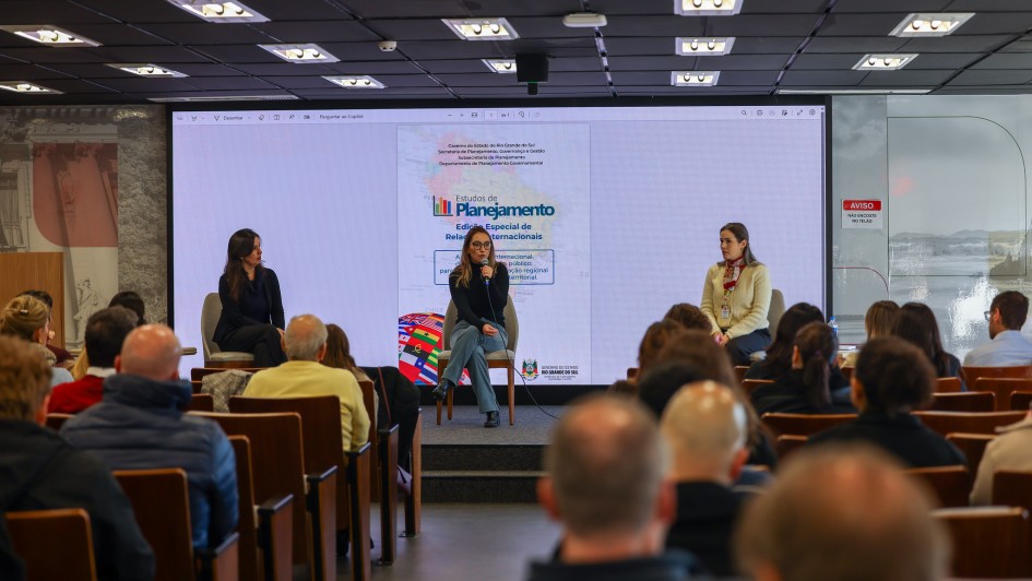A fotografia em plano geral e &acirc;ngulo frontal registra a realiza&ccedil;&atilde;o de um painel de apresenta&ccedil;&atilde;o em um audit&oacute;rio, identificado pelo t&iacute;tulo do arquivo como o lan&ccedil;amento da **Edi&ccedil;&atilde;o especial da revista Estudos de Planejamento apresenta papel das Rela&ccedil;&otilde;es Internacionais no planejamento p&uacute;blico**.

### **O Palco e as Integrantes**

* **A&ccedil;&atilde;o Central:** No centro do palco, uma mulher de &oacute;culos, blusa preta de gola alta e cal&ccedil;a jeans clara est&aacute; sentada em uma cadeira, segurando um microfone e falando ao p&uacute;blico.
* **Demais Painelistas:** Ela &eacute; ladeada por outras duas mulheres sentadas. &Agrave; esquerda, uma mulher veste um conjunto escuro (blazer e cal&ccedil;a); &agrave; direita, outra mulher usa um casaco de tric&ocirc; amarelo-claro com um len&ccedil;o vermelho estampado amarrado ao pesco&ccedil;o e um crach&aacute; institucional.

### **O Painel Digital de Fundo**

* **Proje&ccedil;&atilde;o:** Atr&aacute;s das painelistas, uma grande tela digital estende-se por quase toda a parede do fundo, exibindo a capa da publica&ccedil;&atilde;o t&eacute;cnica em formato digital.
* **Elementos da Capa:** No slide projetado, l&ecirc;-se o t&iacute;tulo em destaque: **"Estudos de Planejamento - Edi&ccedil;&atilde;o Especial de Rela&ccedil;&otilde;es Internacionais"**. O documento traz grafado na parte superior as inst&acirc;ncias institucionais: "Governo do Estado do Rio Grande do Sul / Secretaria de Planejamento, Governan&ccedil;a e Gest&atilde;o / Subsecretaria de Planejamento / Departamento de Planejamento Governamental". Na parte inferior da proje&ccedil;&atilde;o, veem-se ilustra&ccedil;&otilde;es estilizadas de um conjunto de bandeiras de diferentes pa&iacute;ses e o logotipo oficial do governo do estado.

### **O Ambiente e a Plateia**

* **A Plateia:** Em primeiro plano, v&aacute;rias fileiras de poltronas de madeira com encosto castanho est&atilde;o preenchidas por espectadores assistindo &agrave; apresenta&ccedil;&atilde;o, vistos predominantemente de costas.
* **Infraestrutura do Local:** O audit&oacute;rio possui teto rebaixado de placas pretas quadriculadas, equipado com diversas lumin&aacute;rias de LED quadradas embutidas e acesas. &Agrave; direita da tela, h&aacute; uma mureta de vidro com uma placa de sinaliza&ccedil;&atilde;o vertical vermelha e branca onde se l&ecirc; em letras mai&uacute;sculas: **"AVISO / N&Atilde;O ENCOSTE NO TEL&Atilde;O"**.
