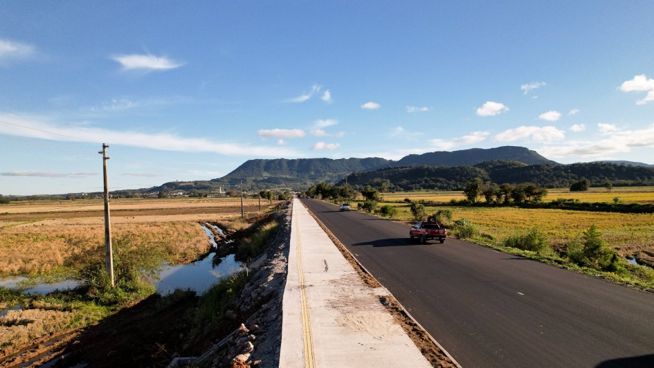 Rodovia asfaltada em &aacute;rea rural plana, com ve&iacute;culos circulando e acostamento claro ao lado, ladeada por canais de &aacute;gua e extensas planta&ccedil;&otilde;es; postes de energia acompanham a via e, ao fundo, h&aacute; morros e c&eacute;u azul com poucas nuvens.
