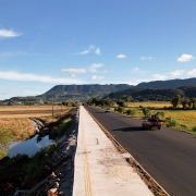 Rodovia asfaltada em &aacute;rea rural plana, com ve&iacute;culos circulando e acostamento claro ao lado, ladeada por canais de &aacute;gua e extensas planta&ccedil;&otilde;es; postes de energia acompanham a via e, ao fundo, h&aacute; morros e c&eacute;u azul com poucas nuvens.
