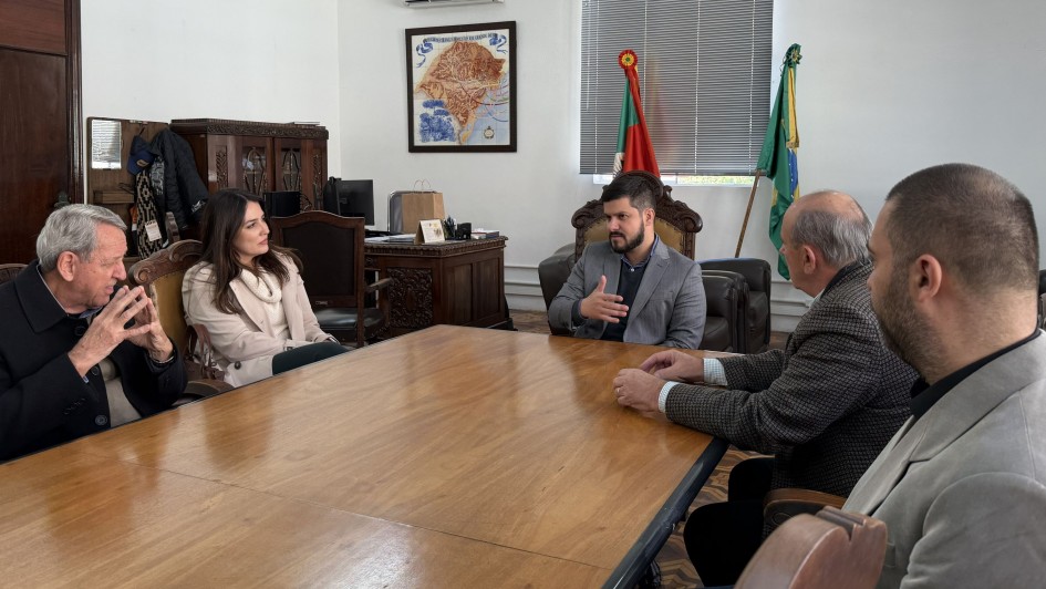A fotografia em plano m&eacute;dio registra uma reuni&atilde;o de trabalho em um gabinete governamental, identificada pelo t&iacute;tulo do arquivo como o ato em que o **"Rio Grande do Sul realiza ato simb&oacute;lico de renova&ccedil;&atilde;o do seguro pecu&aacute;rio ga&uacute;cho contra a febre aftosa.jpg"**.

### **Os Participantes e a Reuni&atilde;o**

* **A Disposi&ccedil;&atilde;o:** Cinco pessoas (quatro homens e uma mulher) est&atilde;o sentadas ao redor de uma grande mesa de reuni&otilde;es retangular de madeira maci&ccedil;a e clara.
* **Lado Esquerdo da Mesa:**
* Em primeiro plano, um homem idoso de cabelos grisalhos e casaco preto gesticula com as duas m&atilde;os unidas pr&oacute;ximas ao rosto enquanto fala.
* Ao seu lado, uma mulher de cabelos longos escuros acompanha a conversa. Ela veste uma blusa branca de gola alta sob um casaco sobretudo claro.


* **Lado Direito e Centro:**
* Na cabeceira ao fundo, centralizado, um homem jovem com barba aparada e vestindo um palet&oacute; cinza sobre camisa escura gesticula com a m&atilde;o direita, conduzindo ou respondendo a uma fala.
* &Agrave; direita da mesa, de perfil parcial para a c&acirc;mera, um homem calvo de cabelos grisalhos veste um palet&oacute; quadriculado (tweed) em tons escuros sobre uma camisa social clara com punhos &agrave; mostra.
* Em primeiro plano, na extrema direita, v&ecirc;-se o perfil da cabe&ccedil;a e ombros de um homem de cabelos curtos escuros vestindo um blazer cinza-claro.



### **O Gabinete e a Decora&ccedil;&atilde;o**

* **Mobili&aacute;rio de &Eacute;poca:** As cadeiras utilizadas possuem encostos altos de madeira escura com entalhes ornamentados cl&aacute;ssicos e assentos estofados. Ao fundo, no lado esquerdo, h&aacute; arm&aacute;rios e escrivaninhas de madeira maci&ccedil;a trabalhada, caracter&iacute;sticos de escrit&oacute;rios institucionais tradicionais.
* **S&iacute;mbolos Oficiais:** Na parede branca ao fundo, centralizado, h&aacute; um quadro com uma moldura escura exibindo um mapa antigo ou tem&aacute;tico do Estado do Rio Grande do Sul. Mais &agrave; direita, posicionadas ao lado de uma janela com persianas cinzas fechadas, destacam-se duas bandeiras oficiais em seus pedestais: a bandeira do Estado do Rio Grande do Sul (com suas listras verde, vermelha e amarela) e a bandeira do Brasil. Um aparelho de ar-condicionado do tipo *split* est&aacute; fixado pr&oacute;ximo ao teto, acima do quadro.
