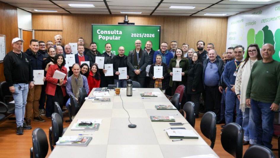 Foto mostra o secret&aacute;rio, os prefeitos e assessores posando para foto em uma sala de reuni&otilde;es. Ao fundo, uma tela verde mostra o nome Consulta Popular .