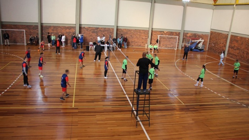 Foto mostra uma quadra de v&ocirc;lei onde acontece uma partida. Um time est&aacute; com camiseta verde claro, outro azul escuro. 