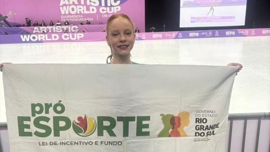 A fotografia em plano inteiro e orienta&ccedil;&atilde;o vertical registra uma jovem patinadora em p&eacute;, segurando uma bandeira institucional em frente a uma pista de patina&ccedil;&atilde;o art&iacute;stica. O t&iacute;tulo do arquivo identifica a atleta como Manuela, vinculada ao programa governamental, sob o nome **"PatinadoraManuelaProEsporteRS.jpg"**.

### **A Atleta e a Bandeira**

* **A Patinadora:** No centro da imagem, uma menina de pele clara, cabelos ruivos presos e um sorriso aberto posa voltada para a c&acirc;mera. Ela usa batom escuro e est&aacute; vestida com trajes de patina&ccedil;&atilde;o, sendo poss&iacute;vel ver suas meias cal&ccedil;as cor da pele cobrindo os p&eacute;s e t&ecirc;nis esportivos nas cores branca, vermelha e rosa na base.
* **A Bandeira:** Com os bra&ccedil;os estendidos para as laterais, ela segura uma grande bandeira branca retangular que cobre a maior parte de seu corpo.
* No lado esquerdo da bandeira, destaca-se o logotipo **"pr&oacute; ESPORTE"** em letras verdes, acompanhado pelo texto menor **"LEI DE INCENTIVO E FUNDO"**.
* No lado direito, h&aacute; silhuetas estilizadas de perfis humanos coloridos e a inscri&ccedil;&atilde;o oficial **"GOVERNO DO ESTADO / RIO GRANDE DO SUL"**, seguida abaixo por "SECRETARIA DO ESPORTE E LAZER".
* Na parte inferior centralizada da bandeira, aparece o logotipo da empresa **"CEGIL / SUPERMERCADOS"** em letras azuis, encimado por um semic&iacute;rculo amarelo e verde.



### **O Cen&aacute;rio e o Ambiente**

* **A Pista de Patina&ccedil;&atilde;o:** Ao fundo, estende-se uma ampla pista de gelo ou piso liso e claro, t&iacute;pica para competi&ccedil;&otilde;es de patina&ccedil;&atilde;o art&iacute;stica. A mureta que delimita a pista exibe banners na cor roxa com a inscri&ccedil;&atilde;o em letras mai&uacute;sculas brancas: **"ARTISTIC WORLD CUP"**.
* **Painel e Tel&atilde;o de Fundo:** Atr&aacute;s da pista, h&aacute; uma grande estrutura cenogr&aacute;fica roxa com as mesmas inscri&ccedil;&otilde;es da competi&ccedil;&atilde;o e indica&ccedil;&otilde;es de locais como "GARMISCH-PARTENKIRCHEN". No canto superior direito, um grande tel&atilde;o digital exibe a transmiss&atilde;o ao vivo ou replays de uma apresenta&ccedil;&atilde;o de patina&ccedil;&atilde;o na pista.
* **Infraestrutura e Primeiro Plano:** A atleta est&aacute; posicionada no topo de uma pequena escada ou arquibancada de concreto. Nas laterais inferiores, veem-se poltronas pl&aacute;sticas azuis de arquibancada e uma grade met&aacute;lica cinza de prote&ccedil;&atilde;o. O teto do complexo esportivo &eacute; composto por uma grande estrutura de vigas de madeira aparentes, equipada com calhas de ventila&ccedil;&atilde;o prateadas e sistemas de ilumina&ccedil;&atilde;o e refletores voltados para a pista.