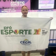 A fotografia em plano inteiro e orienta&ccedil;&atilde;o vertical registra uma jovem patinadora em p&eacute;, segurando uma bandeira institucional em frente a uma pista de patina&ccedil;&atilde;o art&iacute;stica. O t&iacute;tulo do arquivo identifica a atleta como Manuela, vinculada ao programa governamental, sob o nome **"PatinadoraManuelaProEsporteRS.jpg"**.

### **A Atleta e a Bandeira**

* **A Patinadora:** No centro da imagem, uma menina de pele clara, cabelos ruivos presos e um sorriso aberto posa voltada para a c&acirc;mera. Ela usa batom escuro e est&aacute; vestida com trajes de patina&ccedil;&atilde;o, sendo poss&iacute;vel ver suas meias cal&ccedil;as cor da pele cobrindo os p&eacute;s e t&ecirc;nis esportivos nas cores branca, vermelha e rosa na base.
* **A Bandeira:** Com os bra&ccedil;os estendidos para as laterais, ela segura uma grande bandeira branca retangular que cobre a maior parte de seu corpo.
* No lado esquerdo da bandeira, destaca-se o logotipo **"pr&oacute; ESPORTE"** em letras verdes, acompanhado pelo texto menor **"LEI DE INCENTIVO E FUNDO"**.
* No lado direito, h&aacute; silhuetas estilizadas de perfis humanos coloridos e a inscri&ccedil;&atilde;o oficial **"GOVERNO DO ESTADO / RIO GRANDE DO SUL"**, seguida abaixo por "SECRETARIA DO ESPORTE E LAZER".
* Na parte inferior centralizada da bandeira, aparece o logotipo da empresa **"CEGIL / SUPERMERCADOS"** em letras azuis, encimado por um semic&iacute;rculo amarelo e verde.



### **O Cen&aacute;rio e o Ambiente**

* **A Pista de Patina&ccedil;&atilde;o:** Ao fundo, estende-se uma ampla pista de gelo ou piso liso e claro, t&iacute;pica para competi&ccedil;&otilde;es de patina&ccedil;&atilde;o art&iacute;stica. A mureta que delimita a pista exibe banners na cor roxa com a inscri&ccedil;&atilde;o em letras mai&uacute;sculas brancas: **"ARTISTIC WORLD CUP"**.
* **Painel e Tel&atilde;o de Fundo:** Atr&aacute;s da pista, h&aacute; uma grande estrutura cenogr&aacute;fica roxa com as mesmas inscri&ccedil;&otilde;es da competi&ccedil;&atilde;o e indica&ccedil;&otilde;es de locais como "GARMISCH-PARTENKIRCHEN". No canto superior direito, um grande tel&atilde;o digital exibe a transmiss&atilde;o ao vivo ou replays de uma apresenta&ccedil;&atilde;o de patina&ccedil;&atilde;o na pista.
* **Infraestrutura e Primeiro Plano:** A atleta est&aacute; posicionada no topo de uma pequena escada ou arquibancada de concreto. Nas laterais inferiores, veem-se poltronas pl&aacute;sticas azuis de arquibancada e uma grade met&aacute;lica cinza de prote&ccedil;&atilde;o. O teto do complexo esportivo &eacute; composto por uma grande estrutura de vigas de madeira aparentes, equipada com calhas de ventila&ccedil;&atilde;o prateadas e sistemas de ilumina&ccedil;&atilde;o e refletores voltados para a pista.