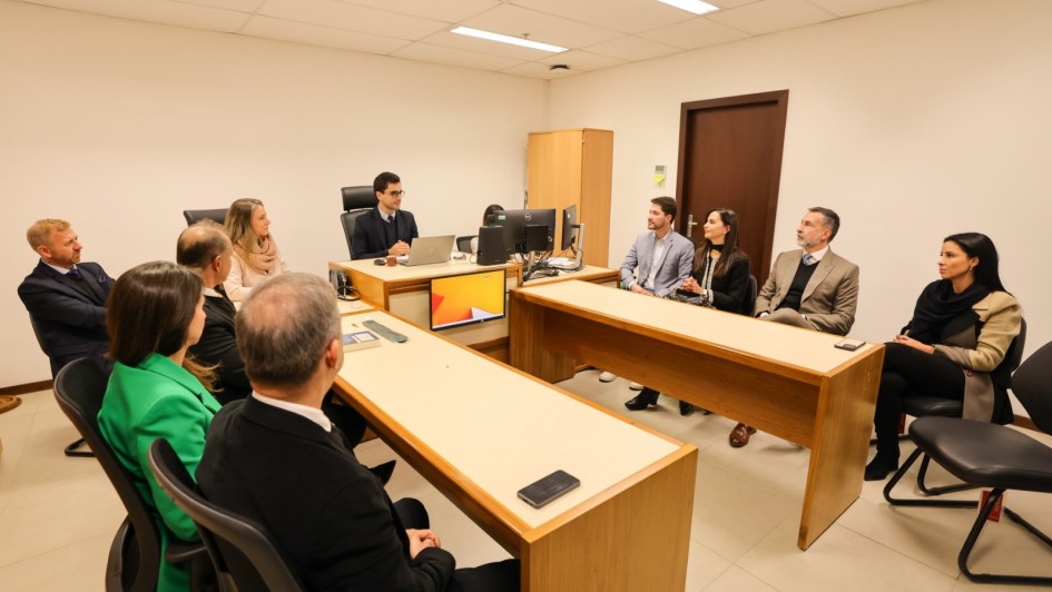 A imagem mostra uma sala de reuni&otilde;es, com pessoas sentadas ao redor de mesas de escrit&oacute;rio. 