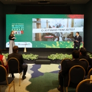 A fotografia em plano geral e &acirc;ngulo frontal registra uma palestra durante um evento institucional em um audit&oacute;rio, identificado pelo t&iacute;tulo do arquivo como **"Foto 1 - Governo do Estado apresenta Projeto Escola &Iacute;ntegra como boa pr&aacute;tica de promo&ccedil;&atilde;o da &eacute;tica em evento nacional.jpg"**.

### **O Palco e os Palestrantes**

* **A Palestrante:** No lado esquerdo do palco, uma mulher com cabelos longos est&aacute; em p&eacute;, segurando um microfone na m&atilde;o direita e folhas de papel na m&atilde;o esquerda. Ela veste uma blusa preta de mangas compridas e uma cal&ccedil;a de alfaiataria cinza de corte largo (pantalona).
* **O Palestrante:** No lado direito do palco, um homem acompanha a apresenta&ccedil;&atilde;o em p&eacute;. Ele veste um terno completo azul-marinho ou preto com gravata escura sobre camisa social clara e segura pap&eacute;is nas m&atilde;os.
* **Mobili&aacute;rio do Palco:** Entre os palestrantes, h&aacute; uma pequena mesa redonda de apoio com base c&ocirc;nica dourada, sobre a qual repousa um copo ou objeto de vidro. A borda frontal do palco &eacute; decorada com uma fileira cont&iacute;nua de folhagens verdes baixas em vasos.

### **O Tel&atilde;o Digital e Conte&uacute;do**

Atr&aacute;s dos palestrantes, um grande tel&atilde;o digital de LED preenche o fundo do palco, dividido visualmente em duas partes sobre um fundo verde texturizado:

* **Lado Esquerdo:** Exibe a marca do evento em letras brancas e amarelas: **"Ciclo de Palestras / 58&ordf; REUNI&Atilde;O T&Eacute;CNICA DO CONACI"**, seguido por quatro pequenas imagens quadradas coloridas e a indica&ccedil;&atilde;o de local: **"PORTO ALEGRE | RS"**. Na borda inferior esquerda, veem-se logotipos de institui&ccedil;&otilde;es organizadoras e parceiras.
* **Lado Direito:** Mostra um slide com uma fotografia retangular centralizada que retrata o corredor de um hospital p&uacute;blico movimentado, com macas e pessoas. Sobreposta a essa foto hospitalar, h&aacute; uma faixa branca horizontal com efeito de papel rasgado onde se l&ecirc; em letras mai&uacute;sculas pretas: **"O JEITINHO TEM JEITO?"**.

### **O Ambiente e a Plateia**

* **Primeiro Plano:** Em primeiro plano, a plateia &eacute; vista de costas, sentada em cadeiras estofadas pretas com estruturas met&aacute;licas douradas, dispostas em fileiras organizadas. Os espectadores, em sua maioria homens e mulheres vestindo trajes formais (blazers e palet&oacute;s), observam a apresenta&ccedil;&atilde;o.
* **Cen&aacute;rio:** O audit&oacute;rio possui paredes laterais em tom verde-escuro opaco. O ch&atilde;o entre o palco e a plateia &eacute; revestido por um tapete com estampas abstratas e org&acirc;nicas em tons de azul, verde, bege e cinza. A ilumina&ccedil;&atilde;o &eacute; focada diretamente sobre o palco e o tel&atilde;o, mantendo a &aacute;rea do p&uacute;blico sob luz mais suave.