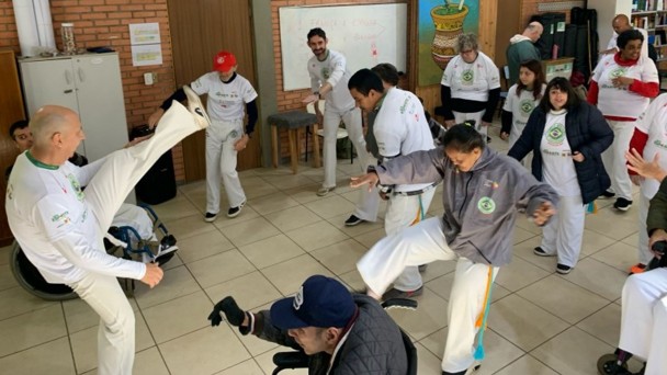 Foto mostra uma aula de capoeira dentro de uma sala de aula. O mestre faz um movimento que &eacute; imitado pelos demais participantes. 