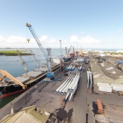 A fotografia a&eacute;rea apresenta uma vista panor&acirc;mica de um porto comercial em plena atividade. O cen&aacute;rio &eacute; dominado por grandes infraestruturas log&iacute;sticas, navios de carga e componentes industriais sob um c&eacute;u claro com algumas nuvens esparsas. 