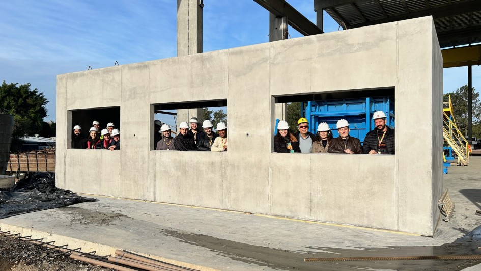 Esta &eacute; uma descri&ccedil;&atilde;o da imagem para fins de acessibilidade:

A fotografia em plano geral e &acirc;ngulo frontal registra a inspe&ccedil;&atilde;o t&eacute;cnica de uma estrutura modular pr&eacute;-moldada de concreto em um canteiro de obras ou f&aacute;brica de elementos construtivos. O t&iacute;tulo do arquivo identifica a a&ccedil;&atilde;o: **"Equipe da SOP vistoria produ&ccedil;&atilde;o dos m&oacute;dulos que integrar&atilde;o nova escola em Gravata&iacute;.jpg"**.

### **A Estrutura de Concreto e a Equipe**

* **O M&oacute;dulo:** No centro do plano, destaca-se uma grande estrutura retangular e maci&ccedil;a de concreto cinza-claro, com textura fosca e lisa. A parede frontal da estrutura possui tr&ecirc;s grandes aberturas retangulares sim&eacute;tricas que servir&atilde;o como janelas.
* **A Equipe de Vistoria:** Uma equipe de aproximadamente 15 profissionais est&aacute; posicionada lado a lado por tr&aacute;s da parede de concreto, espiando e posando para a foto atrav&eacute;s das tr&ecirc;s aberturas das janelas. Todos usam crach&aacute;s institucionais pendurados no pesco&ccedil;o e capacetes de prote&ccedil;&atilde;o (EPIs), a grande maioria na cor branca. Eles sorriem em dire&ccedil;&atilde;o &agrave; c&acirc;mera.
* **Na janela da esquerda:** Um grupo de cerca de cinco pessoas est&aacute; reunido, vestindo casacos escuros.
* **Na janela do centro:** Outro grupo de cinco profissionais, incluindo homens com jaquetas escuras e camisas claras.
* **Na janela da direita:** Um grupo de cinco pessoas est&aacute; perfilado. Entre elas, destaca-se um homem usando &oacute;culos escuros e capacete amarelo, ladeado por colegas vestindo casacos de inverno (incluindo uma jaqueta de couro marrom e um casaco acolchoado preto na extrema direita).



### **O Entorno da F&aacute;brica e Constru&ccedil;&atilde;o**

* **O Ch&atilde;o:** A estrutura modular est&aacute; assentada sobre uma base plana de concreto claro. &Agrave; frente dela, no quadrante inferior esquerdo, h&aacute; uma lona pl&aacute;stica preta estendida no ch&atilde;o e barras met&aacute;licas de arma&ccedil;&atilde;o (vergalh&otilde;es) dispostas horizontalmente pr&oacute;ximas &agrave; borda da cal&ccedil;ada.
* **A Infraestrutura ao Fundo:** O local &eacute; amplo e parcialmente coberto. Acima do m&oacute;dulo de concreto, erguem-se altas colunas e vigas estruturais de concreto que sustentam uma cobertura met&aacute;lica industrial vis&iacute;vel no canto superior direito. Atr&aacute;s da abertura da janela direita, avista-se uma grande f&ocirc;rma ou maquin&aacute;rio industrial na cor azul-viva. Na extrema direita, ao fundo, nota-se uma escada ou passarela met&aacute;lica amarela de acesso t&eacute;cnico.
* **C&eacute;u e Ilumina&ccedil;&atilde;o:** O dia est&aacute; claro e ensolarado, com o c&eacute;u azul e poucas nuvens esparsas vis&iacute;veis no topo esquerdo. A ilumina&ccedil;&atilde;o natural projeta sombras suaves no ch&atilde;o &agrave; frente do m&oacute;dulo.