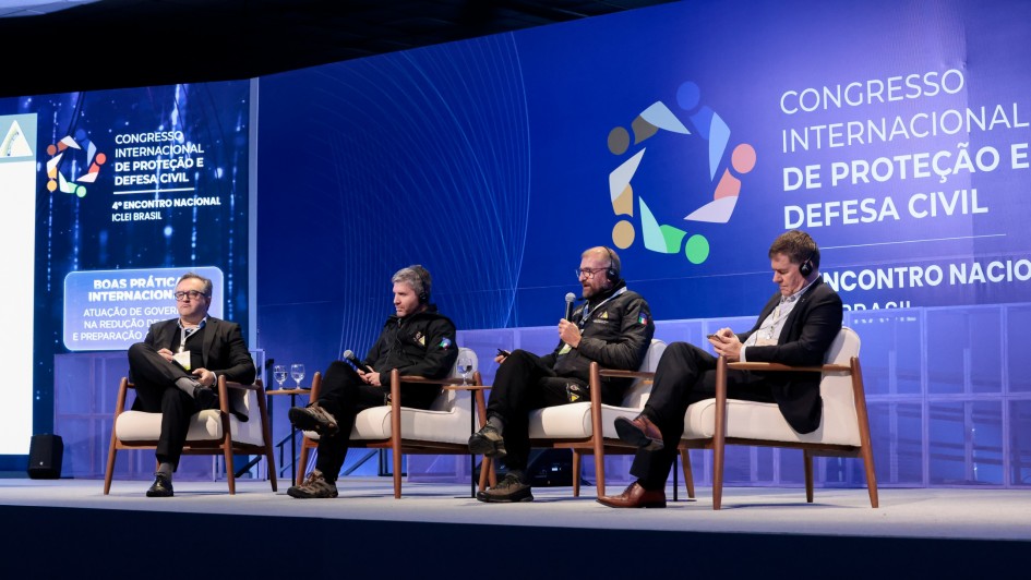 Quatro pessoas est&atilde;o em um palco, sentadas em cadeiras, uma delas fala ao microfone. Atr&aacute;s, um grande painel indicando Congresso Internacional de Prote&ccedil;&atilde;o e Defesa Civil