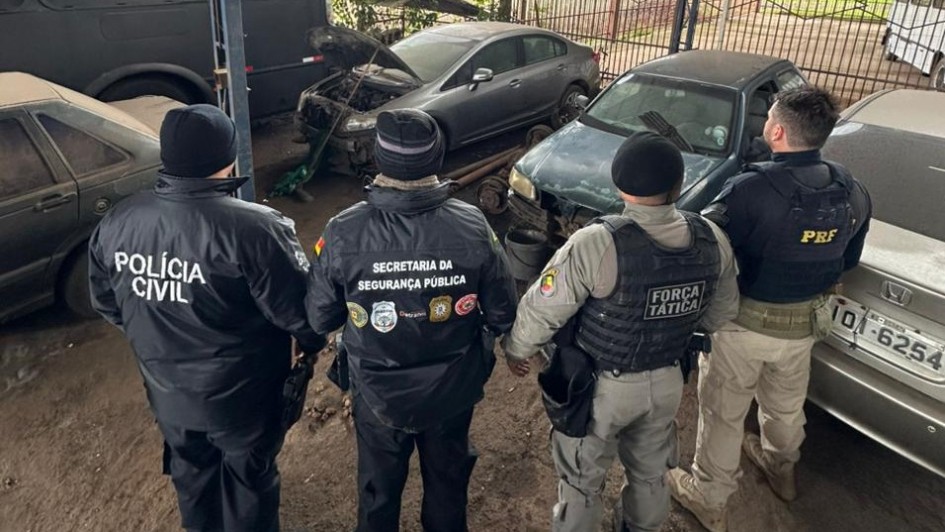 Quatro policiais est&atilde;o parados em frente a v&aacute;rias sucatas de ve&iacute;culos em um p&aacute;tio de desmanche