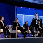 Esta &eacute; uma descri&ccedil;&atilde;o da imagem para fins de acessibilidade:

A fotografia em plano m&eacute;dio e &acirc;ngulo frontal-lateral registra a realiza&ccedil;&atilde;o de um painel de debates sobre um palco de eventos, identificado pelo t&iacute;tulo do arquivo como **"Painel Boas Praticas - foto Jo&atilde;o Pedro Rodrigues.jpg"**.

### **Os Integrantes do Painel**

* **Composi&ccedil;&atilde;o:** Quatro painelistas est&atilde;o sentados em uma fileira de poltronas brancas ou creme individuais com bra&ccedil;os e pernas de madeira clara, dispostas ao longo do palco. Todos usam credenciais oficiais suspensas por cord&otilde;es no pesco&ccedil;o.
* **Da esquerda para a direita:**
* Uma mulher de &oacute;culos e cabelos cacheados escuros, vestindo casaco preto e botas marrons de salto, est&aacute; sentada voltada de perfil parcial para a direita, segurando um microfone na m&atilde;o esquerda.
* Uma mulher de cabelos longos loiros ou claros est&aacute; centralizada no palco, falando ao microfone que segura com a m&atilde;o direita. Ela veste um casaco preto, cal&ccedil;a escura e botas pretas de salto.
* Uma mulher de &oacute;culos e cabelos curtos escuros est&aacute; sentada em pose reflexiva, acompanhando a fala da colega. Ela veste um casaco escuro de inverno e sapatos pretos.
* Um homem jovem de cabelos escuros e curtos est&aacute; sentado na extremidade direita do grupo, vestindo um su&eacute;ter preto sobre camisa social e cal&ccedil;a escura, com as m&atilde;os unidas sobre as pernas.


* **Extrema Esquerda:** Na borda esquerda inferior da imagem, veem-se apenas as pernas de outra participante vestindo cal&ccedil;a escura e botas de couro pretas de cano alto com fivelas douradas.

### **O Tel&atilde;o e o Cen&aacute;rio de Fundo**

* **O Grande Painel de LED:** Atr&aacute;s dos participantes, estende-se um imenso tel&atilde;o digital com ilumina&ccedil;&atilde;o e fundo predominantemente na cor azul-viva.
* **Conte&uacute;do Digital:** O lado esquerdo do tel&atilde;o exibe grafismos abstratos compostos por linhas finas e curvas que se cruzam de forma ritmada. O lado direito projeta em tamanho ampliado a transmiss&atilde;o ao vivo em v&iacute;deo (transmiss&atilde;o simult&acirc;nea) da painelista loira central enquanto ela realiza o seu pronunciamento.
* **Mobili&aacute;rio de Apoio:** Pequenas mesas redondas de madeira escura est&atilde;o posicionadas nas laterais e entre as poltronas, servindo de apoio para copos e ta&ccedil;as de &aacute;gua de vidro transparente. O teto do audit&oacute;rio, com placas quadradas escuras e sistemas de ilumina&ccedil;&atilde;o embutidos, aparece de forma discreta no topo da composi&ccedil;&atilde;o.
