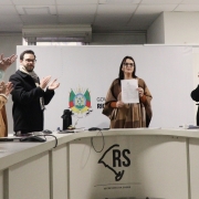 A fotografia em plano geral e &acirc;ngulo frontal registra o momento posterior &agrave; assinatura de um ato oficial em uma sala de reuni&otilde;es governamental. O t&iacute;tulo do arquivo identifica o contexto como a **"Assinatura da portaria que instituiu a Pol&iacute;tica Estadual de Monitoramento e Avalia&ccedil;&atilde;o em Sa&uacute;de 2026.jpeg"**.

### **As Autoridades e a A&ccedil;&atilde;o Central**

* **A Autoridade Central:** No centro do plano, atr&aacute;s de uma grande bancada, uma mulher de cabelos escuros e &oacute;culos de grau est&aacute; em p&eacute;, voltada para a frente. Ela segura com a m&atilde;o esquerda um documento oficial impresso em folha branca, exibindo-o para a c&acirc;mera. Ela veste uma blusa terracota sob um poncho de l&atilde; em tons de marrom e bege com estampa quadriculada.
* **Grupo de Apoio (&agrave; esquerda):**
* Um homem de &oacute;culos, barba e vestindo um casaco preto espesso com um cachecol de padr&atilde;o xadrez claro est&aacute; em p&eacute;, sorrindo e batendo palmas em dire&ccedil;&atilde;o &agrave; autoridade central.
* Ao lado dele, mais &agrave; esquerda, duas mulheres tamb&eacute;m aparecem em p&eacute; batendo palmas; uma veste um casaco caramelo e a outra, mais pr&oacute;xima &agrave; borda esquerda, veste um su&eacute;ter de tric&ocirc; verde-claro. Uma m&atilde;o masculina com casaco escuro aparece no canto inferior esquerdo segurando uma caneta.


* **Autoridade &agrave; Direita:** No lado direito da imagem, outra mulher de &oacute;culos com arma&ccedil;&atilde;o avermelhada e cabelos presos acompanha o ato em p&eacute;. Ela sorri e bate palmas, vestindo um casaco preto de inverno sobre uma echarpe ou blusa rosa vibrante.

### **O Ambiente e Elementos Institucionais**

* **A Bancada:** As autoridades est&atilde;o posicionadas atr&aacute;s de uma grande mesa ou bancada curva de trabalho em tom cinza-claro ou branco. Na face frontal dessa bancada, voltada para o espectador, destaca-se a aplica&ccedil;&atilde;o em preto do logotipo oficial estilizado do mapa do Rio Grande do Sul acompanhado da inscri&ccedil;&atilde;o **"RS / SECRETARIA DA SA&Uacute;DE"**. Sobre a mesa, veem-se x&iacute;caras de caf&eacute; brancas, pap&eacute;is, canetas, um mouse e uma garrafa t&eacute;rmica preta com uma cuia de chimarr&atilde;o ao centro.
* **O Painel de Fundo:** Atr&aacute;s das autoridades, h&aacute; um painel ou divis&oacute;ria branca port&aacute;til onde est&aacute; impresso o bras&atilde;o oficial do Estado do Rio Grande do Sul colorido, seguido pelas letras mai&uacute;sculas pretas **"GOV / RIO"** (com o restante do texto encoberto).
* **Cen&aacute;rio:** A sala possui teto de placas modulares brancas com lumin&aacute;rias embutidas e um aparelho de ar-condicionado instalado ao fundo. A ilumina&ccedil;&atilde;o interna &eacute; difusa e uniforme, concentrando a aten&ccedil;&atilde;o no grupo reunido para a celebra&ccedil;&atilde;o do ato.