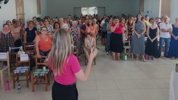 Foto mostra palestrante conduzindo atividade no sal&atilde;o cheio de mulheres. Todas est&atilde;o de p&eacute;.