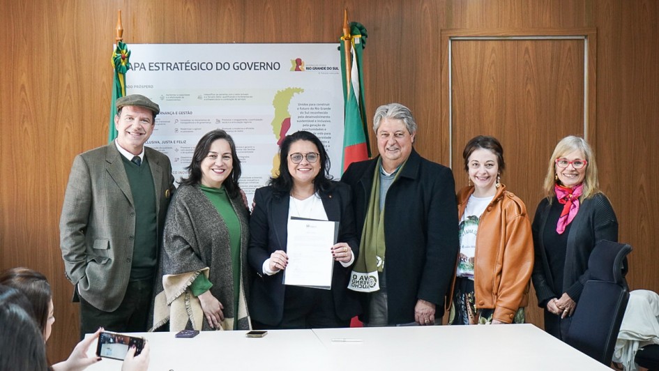 Um grupo de pessoas se posiciona em p&eacute; atr&aacute;s da mesa para uma foto, enquanto a titular da Sema segura um documento voltado para a c&acirc;mera.