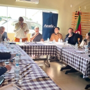 Esta &eacute; uma descri&ccedil;&atilde;o da imagem para fins de acessibilidade:

A fotografia em plano geral e &acirc;ngulo diagonal registra uma reuni&atilde;o de trabalho e articula&ccedil;&atilde;o pol&iacute;tica em um ambiente institucional semi-formal. O t&iacute;tulo do arquivo identifica o evento como **"Governo do Estado refor&ccedil;a parceria com munic&iacute;pios da Zona Sul para ampliar a&ccedil;&otilde;es de enfrentamento &agrave; viol&ecirc;ncia contra as mulheres.jpeg"**.

### **A Oradora e a Mesa Central**

* **O Pronunciamento:** No lado esquerdo do plano de fundo, uma mulher com cabelos curtos loiros ou platinados e &oacute;culos de grau escuros est&aacute; em p&eacute;, falando ao microfone que segura com a m&atilde;o direita. Ela veste um casaco tipo *biker* na cor creme ou esbranqui&ccedil;ada sobre uma blusa clara e cal&ccedil;a ou saia na cor laranja viva.
* **Participantes &agrave; Mesa de Fundo:** Sentados ao longo da extens&atilde;o da mesa ao fundo, acompanhando a fala, est&atilde;o:
* &Agrave; esquerda da oradora, um homem idoso de &oacute;culos e palet&oacute; cinza, inclinado para baixo analisando documentos.
* &Agrave; direita da oradora, um homem de camiseta cinza e &oacute;culos, com a m&atilde;o direita pr&oacute;xima &agrave; boca; ao seu lado, um homem de &oacute;culos e su&eacute;ter marrom-claro com um cord&atilde;o de credencial colorido no pesco&ccedil;o; e mais &agrave; direita, um homem de casaco escuro observando atentamente a oradora.



### **Demais Integrantes e Disposi&ccedil;&atilde;o**

* **Formato da Mesa:** Os participantes est&atilde;o reunidos em torno de uma grande mesa disposta em formato de "U", totalmente coberta por uma toalha com estampa xadrez em tons de marrom-escuro e branco.
* **Ala Direita:** Sentadas no bra&ccedil;o direito da mesa, veem-se uma mulher com cabelos longos escuros voltada em dire&ccedil;&atilde;o &agrave; oradora e, ao seu lado, na extrema direita, um homem sorridente vestindo casaco preto de gola alta.
* **Ala Esquerda (Primeiro Plano):** No bra&ccedil;o esquerdo da mesa, em primeiro plano, aparecem homens sentados de perfil ou de costas para a c&acirc;mera, concentrados na reuni&atilde;o. Um deles, no canto inferior esquerdo, segura um smartphone na m&atilde;o, enquanto os outros revisam pap&eacute;is e relat&oacute;rios distribu&iacute;dos sobre a mesa.
* **Objetos sobre a Mesa:** Ao longo de toda a extens&atilde;o da mesa, encontram-se espalhadas diversas garrafas pl&aacute;sticas transparentes de &aacute;gua mineral, copos de vidro vazios, canetas e microfones de m&atilde;o extras deitados sobre os pap&eacute;is.

### **O Ambiente e S&iacute;mbolos Institucionais**

* **Elementos de Identifica&ccedil;&atilde;o:** Ao fundo, centralizado atr&aacute;s da mesa principal, destaca-se um banner institucional do tipo *roll-up* na cor azul-escura com o logotipo e a inscri&ccedil;&atilde;o: **"Azonasul - Associa&ccedil;&atilde;o dos Munic&iacute;pios da Zona Sul"**.
* **S&iacute;mbolos Oficiais:** No canto superior direito, dispostas em mastros de madeira &agrave; frente de um painel de ripas decorativas de madeira horizontal, encontram-se duas bandeiras oficiais: &agrave; esquerda, a bandeira do Estado do Rio Grande do Sul (com suas faixas verde, vermelha e amarela); &agrave; direita, uma bandeira inteiramente verde adornada com uma roseta de fitas amarela e verde no topo do mastro.
* **Ilumina&ccedil;&atilde;o e Cen&aacute;rio:** O sal&atilde;o possui paredes lisas em tom bege ou ocre claro. Uma grande janela de vidro ao fundo permite a entrada de luz natural abundante, revelando uma fra&ccedil;&atilde;o de carros estacionados do lado de fora. Um aparelho de ar-condicionado *split* branco est&aacute; instalado no alto da parede, logo acima da janela.