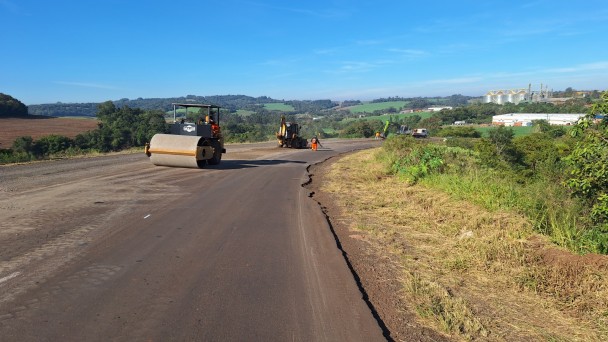 Rodovia em &aacute;rea rural durante obras de pavimenta&ccedil;&atilde;o, com um rolo compactador em primeiro plano e outras m&aacute;quinas e trabalhadores distribu&iacute;dos ao longo da pista; as margens t&ecirc;m vegeta&ccedil;&atilde;o e campos agr&iacute;colas, com morros e silos ao fundo sob c&eacute;u azul aberto.