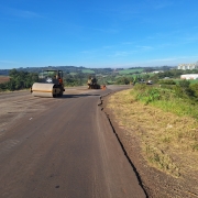 Rodovia em &aacute;rea rural durante obras de pavimenta&ccedil;&atilde;o, com um rolo compactador em primeiro plano e outras m&aacute;quinas e trabalhadores distribu&iacute;dos ao longo da pista; as margens t&ecirc;m vegeta&ccedil;&atilde;o e campos agr&iacute;colas, com morros e silos ao fundo sob c&eacute;u azul aberto.