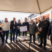 Esta &eacute; uma descri&ccedil;&atilde;o da imagem para fins de acessibilidade:

A fotografia em plano geral e &acirc;ngulo frontal registra o momento posterior &agrave; assinatura de um ato oficial ao ar livre. O t&iacute;tulo do arquivo contextualiza o evento: **"Governo do Estado assina conv&ecirc;nio para duplica&ccedil;&atilde;o da ERS-030 em Os&oacute;rio.jpg"**.

### **As Autoridades e a A&ccedil;&atilde;o Central**

* **O Grupo Central:** No centro da imagem, sob a cobertura de uma tenda, um grupo de autoridades pol&iacute;ticas e representantes p&uacute;blicos est&aacute; perfilado lado a lado, sorrindo em dire&ccedil;&atilde;o &agrave; c&acirc;mera.
* **Exibi&ccedil;&atilde;o do Documento:** O governador do estado est&aacute; posicionado ao centro-direita do grupo, vestindo uma jaqueta escura sobre camiseta escura. Ele segura, junto com o homem de jaqueta azul-marinho &agrave; sua esquerda, uma pasta branca aberta contendo o documento oficial assinado, exibindo-o frontalmente.
* **Demais Integrantes:**
* &Agrave; esquerda do homem de jaqueta azul, um homem de jaqueta preta aberta sobre camisa social clara estende o bra&ccedil;o em dire&ccedil;&atilde;o ao documento. Ao lado deste, um homem de jaqueta bege clara e cal&ccedil;a jeans acompanha o ato sorridente. Na extrema esquerda, veem-se uma mulher com casaco sobretudo claro e outras pessoas ao fundo.
* &Agrave; direita do governador, um homem idoso de &oacute;culos usa uma jaqueta acolchoada cinza escuro. Ao seu lado, destacam-se um homem calvo de jaqueta preta esportiva com os bra&ccedil;os cruzados &agrave; frente e, na extrema direita em primeiro plano, um homem de &oacute;culos e cabelos grisalhos vestindo um su&eacute;ter texturizado azul-marinho.



### **O Mobili&aacute;rio e o Ambiente**

* **A Mesa de Assinatura:** Centralizada em primeiro plano, logo &agrave; frente das autoridades, h&aacute; uma pequena mesa retangular simples com tampo claro e estrutura de metal preto, onde repousa uma caneta escura utilizada para as assinaturas.
* **O Cen&aacute;rio ao Redor:** O grupo est&aacute; posicionado sobre um terreno de terra batida e brita. Eles est&atilde;o abrigados sob uma grande tenda de lona clara, cuja estrutura met&aacute;lica e cobertura aparecem no topo da imagem.
* **O Plano de Fundo:** Ao fundo, sob a luz forte do sol, avistam-se edif&iacute;cios urbanos, incluindo um pr&eacute;dio de dois pavimentos com sacada de grade branca e uma parede de tijolos sem reboco &agrave; direita. Elementos como postes de energia com fia&ccedil;&atilde;o a&eacute;rea e a silhueta de morros distantes na extrema esquerda completam a paisagem externa.